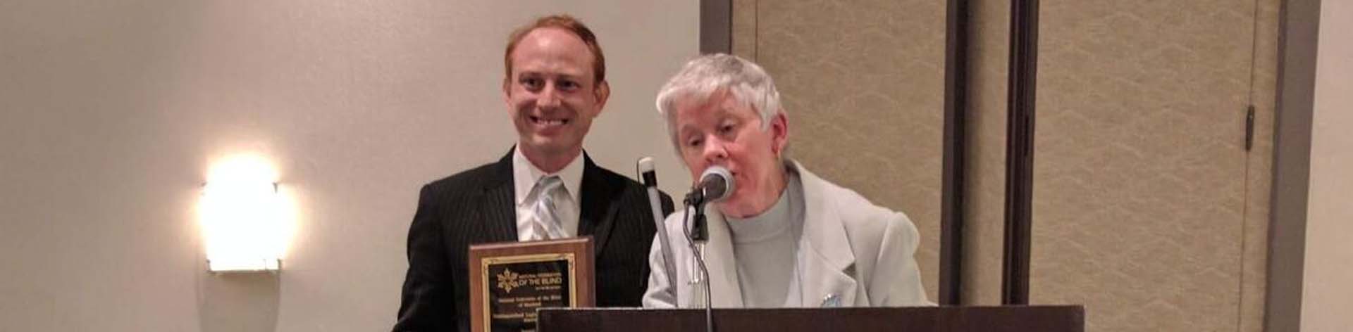 Sharon Maneki speaking at the podium with an award winner holding his plaque in the background