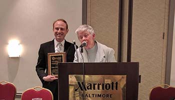 Sharon Maneki speaking at the podium with an award winner holding his plaque in the background