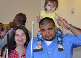 A blind mom and dad smile together. Dad holds their small daughter on his shoulders.